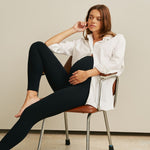 Woman sitting on a chair wearing a white shirt and black leggings against a plain background