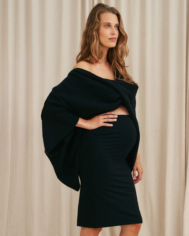 Woman wearing a black off-shoulder top and skirt with high heels against a beige curtain background