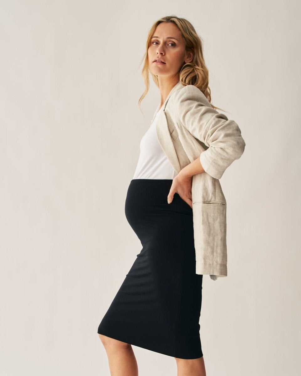 A pregnant woman modeling a black pencil skirt with a beige blazer. The skirt has a stretchy waistband and the woman is standing against a neutral background.