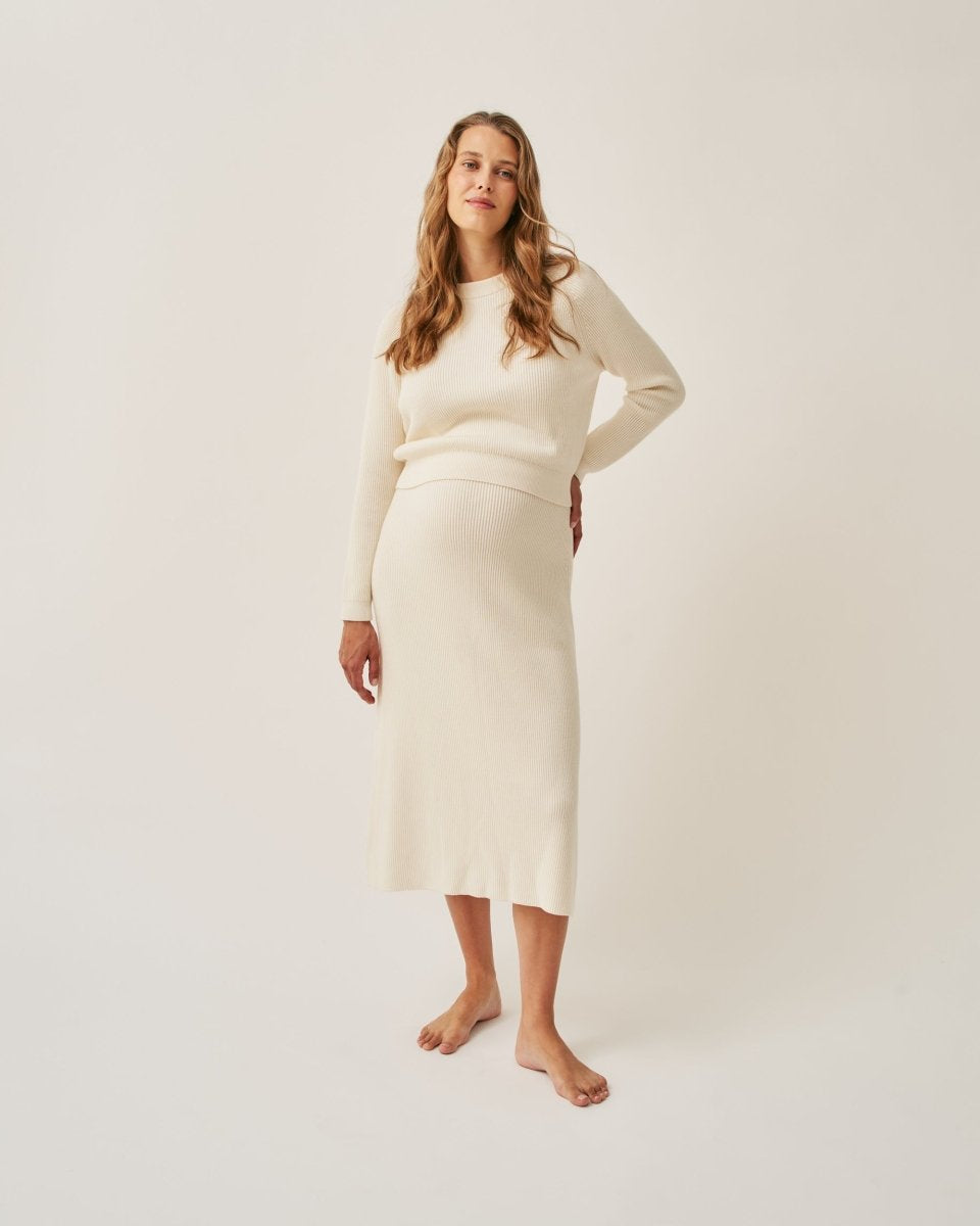 A woman standing in a studio setting, wearing a midi knit skirt in an ecru shade, paired with a matching knit top. The skirt is A-line and the woman is barefoot.