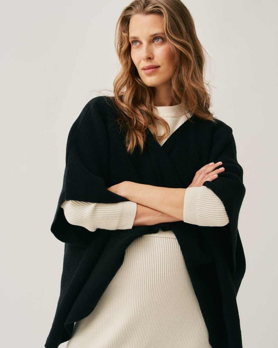 A woman wearing a black poncho with a twist front feature, over a white outfit, standing with her arms crossed.